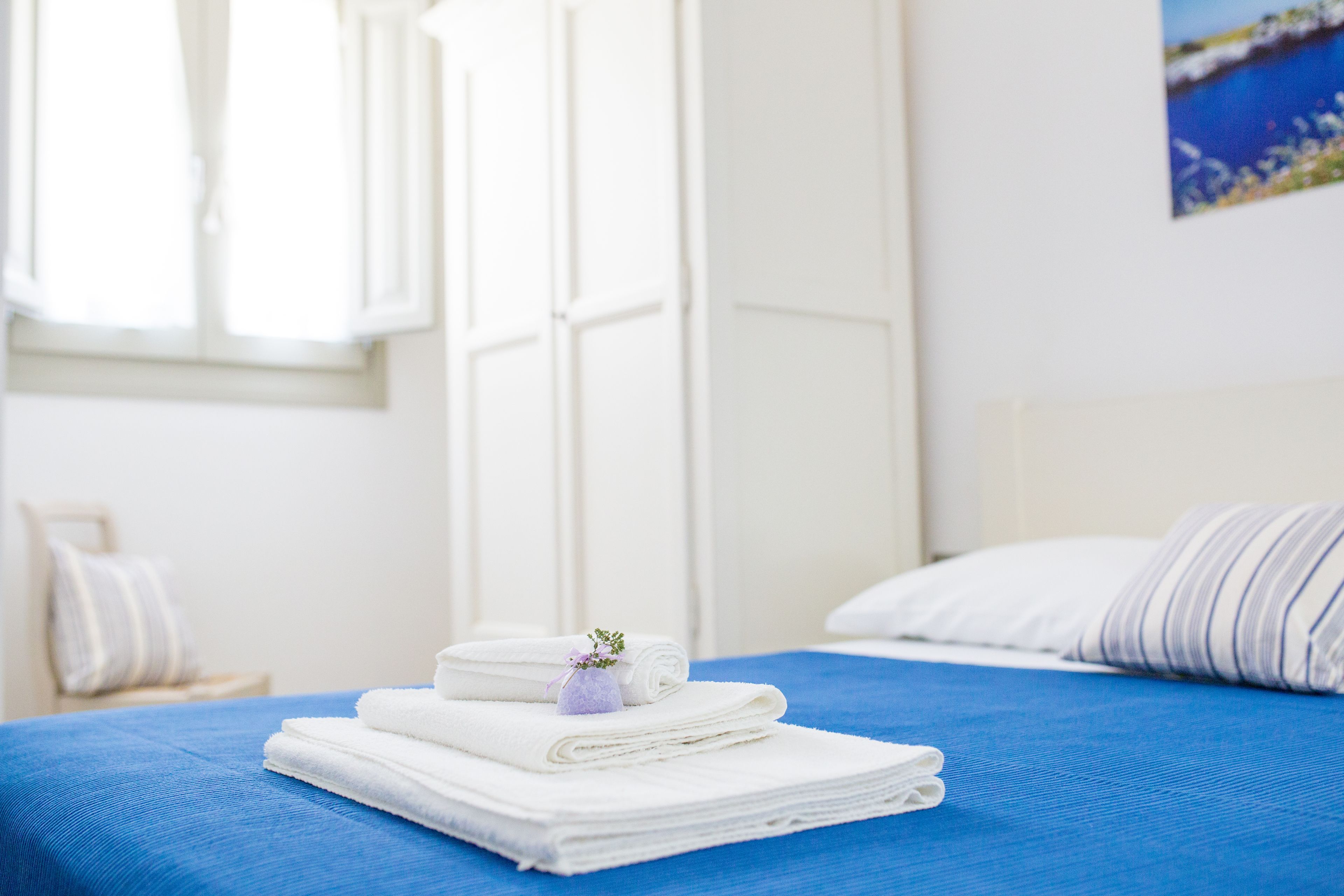 Una cama bien hecha con toallas blancas y una flor morada, una colcha azul y un cabecero blanco.