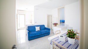 A bright living room with a blue sofa, a dining table with a potted plant, and a view of a balcony with a blue chair. - La Torre nel Borgo (Melendugno)