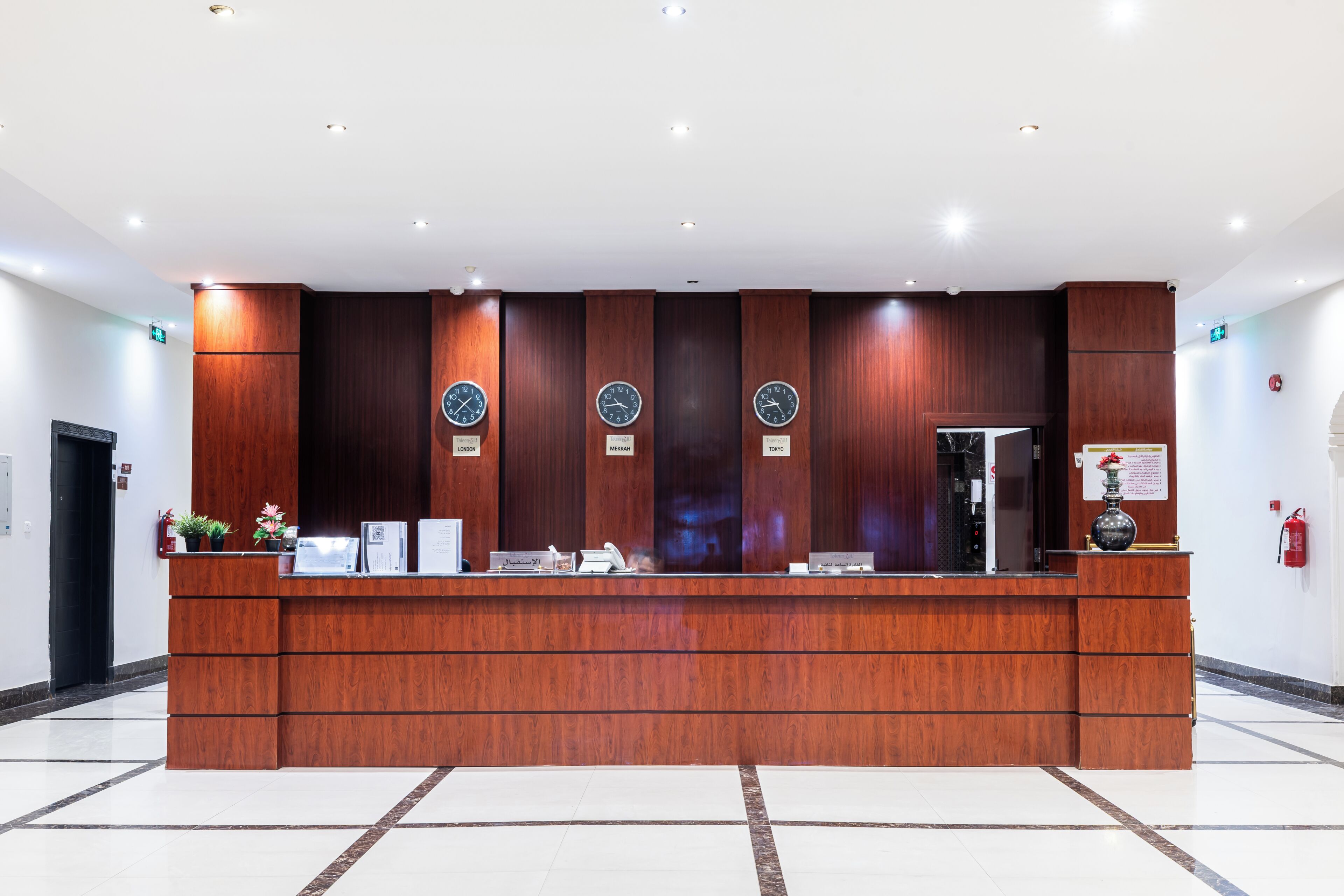 A reception desk with a wooden finish, multiple clocks, a fire extinguisher, and a vase on the right side.