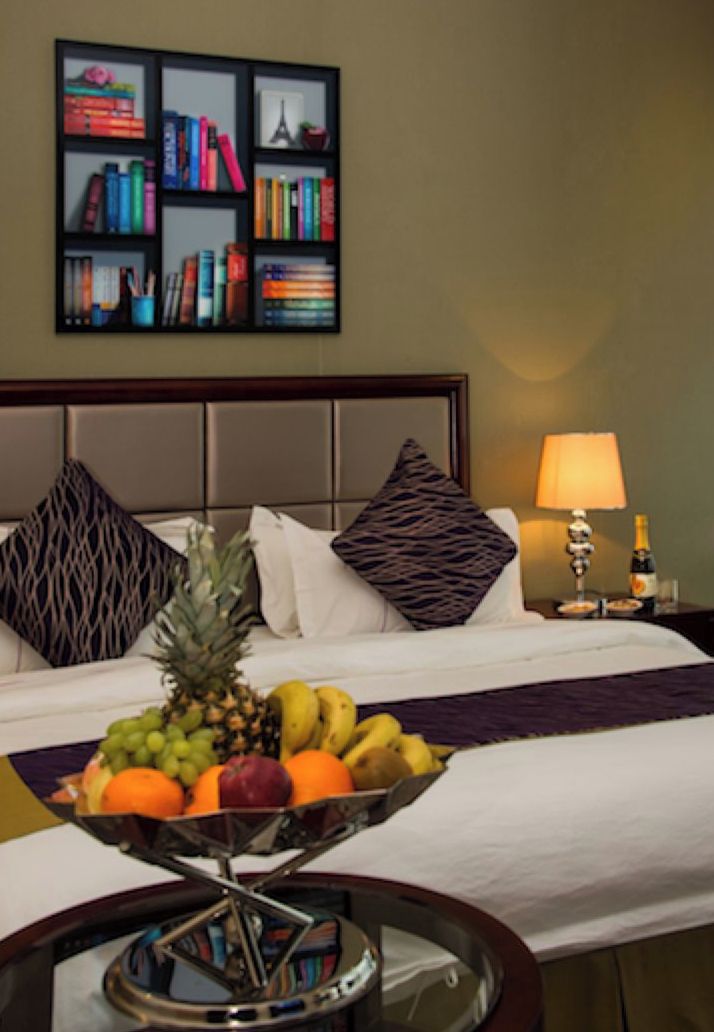 A hotel room with a bed, a bedside table with a lamp, a fruit bowl, and a bookshelf with books and decorative items.
