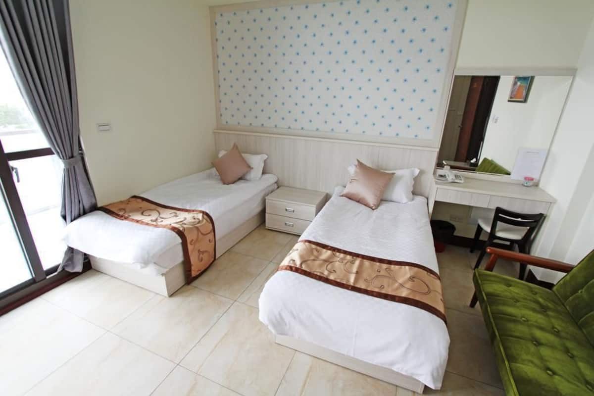 A hotel room with two beds, a desk, a chair, and a large window with curtains.