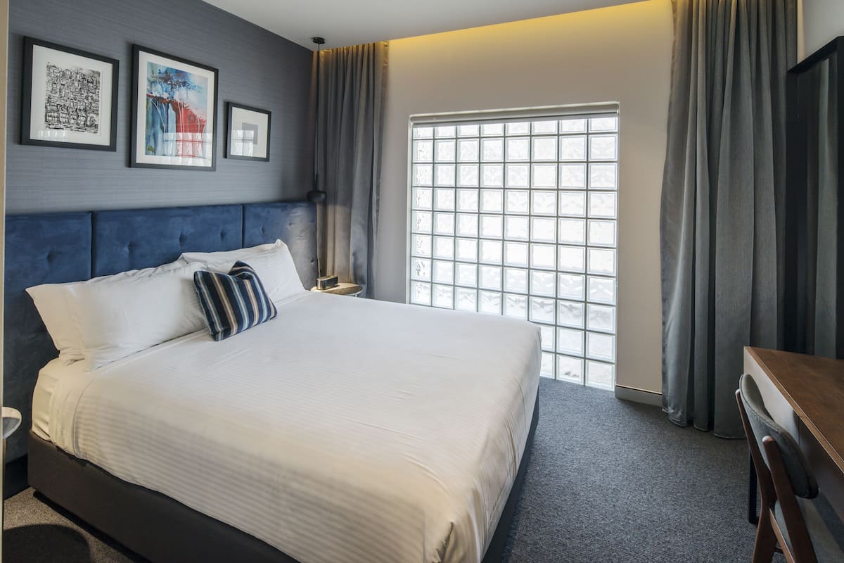 Een net opgemaakt bed met witte lakens en gestreepte kussens, een donkerblauwe hoofdbord en een groot raam met uitzicht op de omgeving.