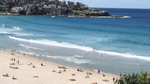 Ein Strand mit Menschen, die sonnenbaden und schwimmen, eine felsige Küste und Gebäude im Hintergrund.