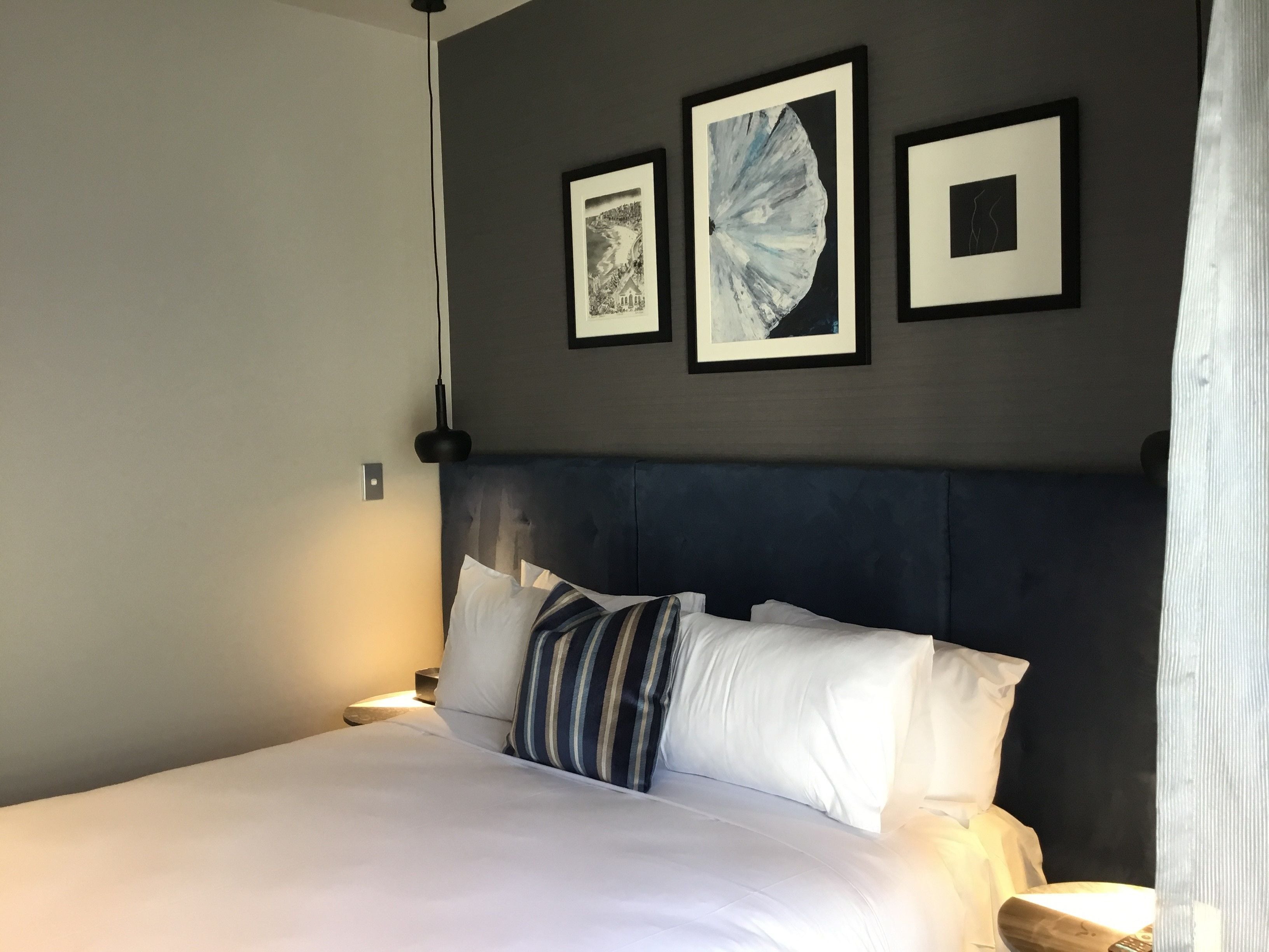 A neatly made bed with white linens and striped pillows, a dark headboard, and three framed artworks on the wall.