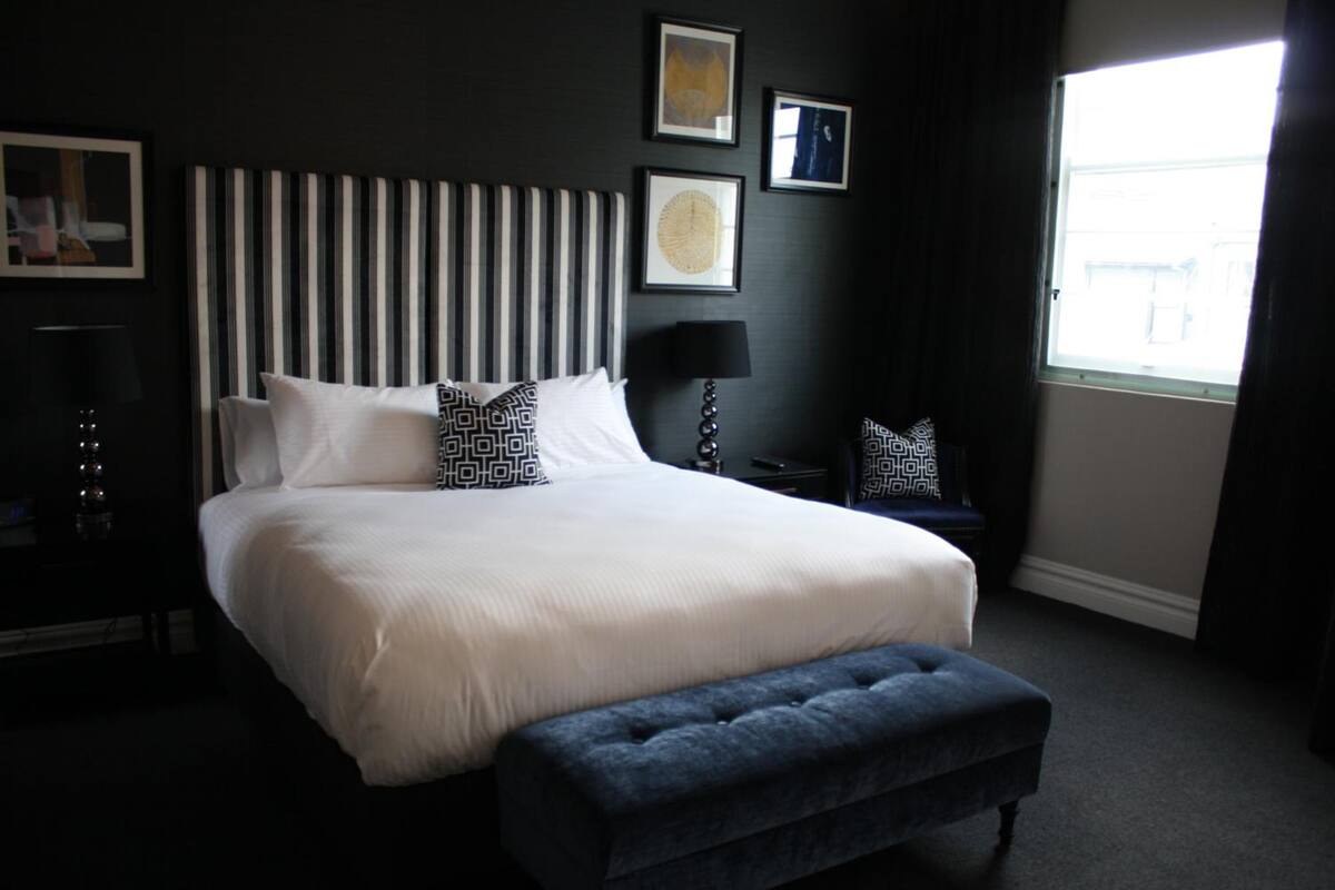 Een keurig opgemaakte slaapkamer met een groot bed, een nachtkastje met een lamp, en een raam dat natuurlijk licht binnenlaat.