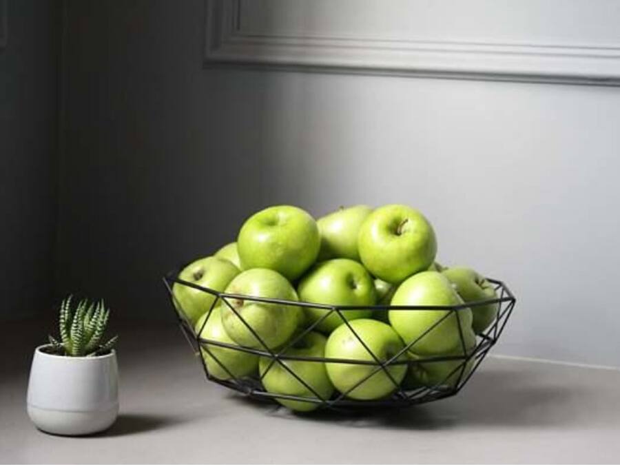 A metal wire basket filled with green apples, a small potted plant, and a grey wall with a door in the background.
