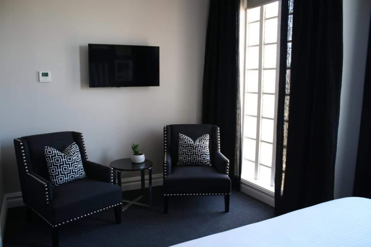 Een hotelkamer met twee fauteuils, een bijzettafeltje met een plant, een flatscreen televisie aan de muur en een groot raam met donkere gordijnen.