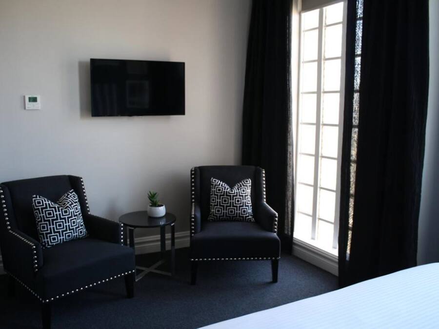 Ein Hotelzimmer mit zwei Sesseln, einem kleinen Tisch mit Pflanze, einem flachbildfernseher an der Wand und einem großen Fenster mit schwarzen Vorhängen.