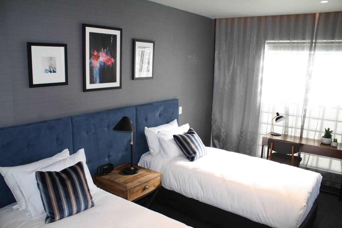 Een hotelkamer met twee bedden, een bureau en een groot raam met gordijnen.
