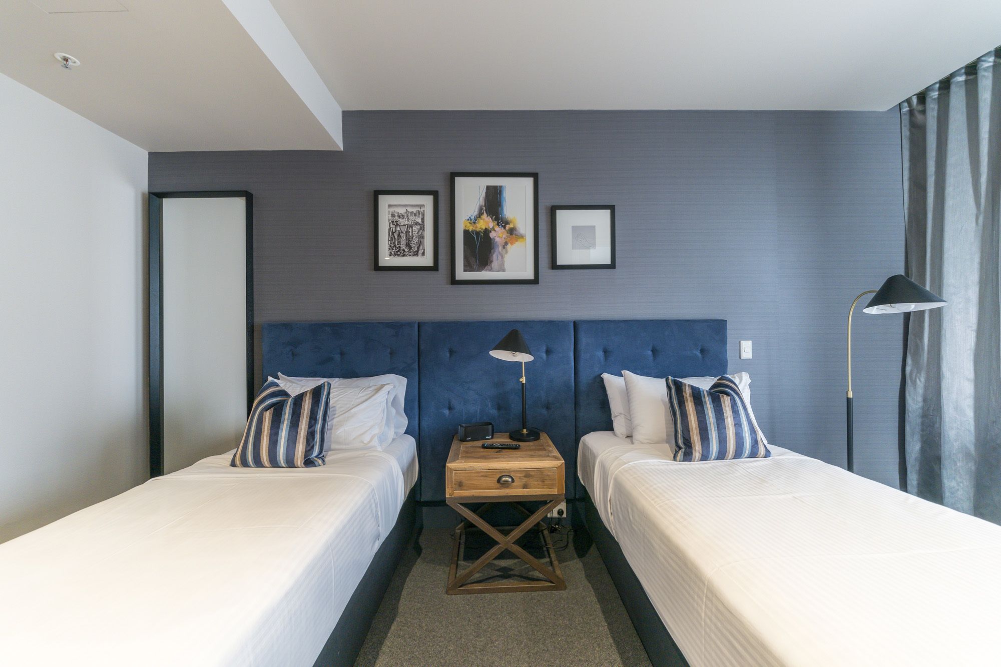 A hotel room with two beds, a nightstand, a lamp, and three framed pictures on the wall.