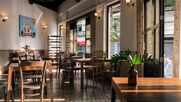 A restaurant interior with wooden tables and chairs, a patterned floor, and a large window with a view of the street. - 103 - Bed and Brews (Bangkok)