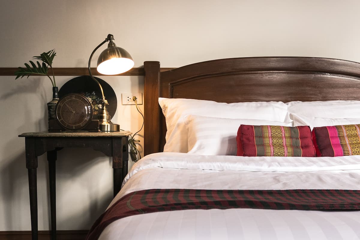 A neatly made bed with white bedding and a striped blanket, a wooden nightstand with a lamp, and a wall clock.