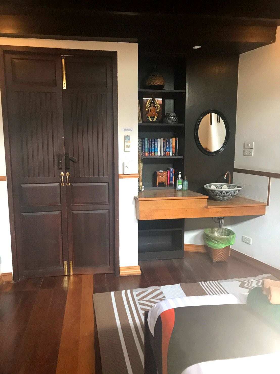 A bedroom with a dark wooden door, a wooden bedside table, a mirror, a shelf with books and decorative items, and a green trash bin.