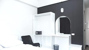 Una habitación de hotel moderna con una cama, una silla negra, un escritorio blanco con un espejo y un cubo de basura negro.