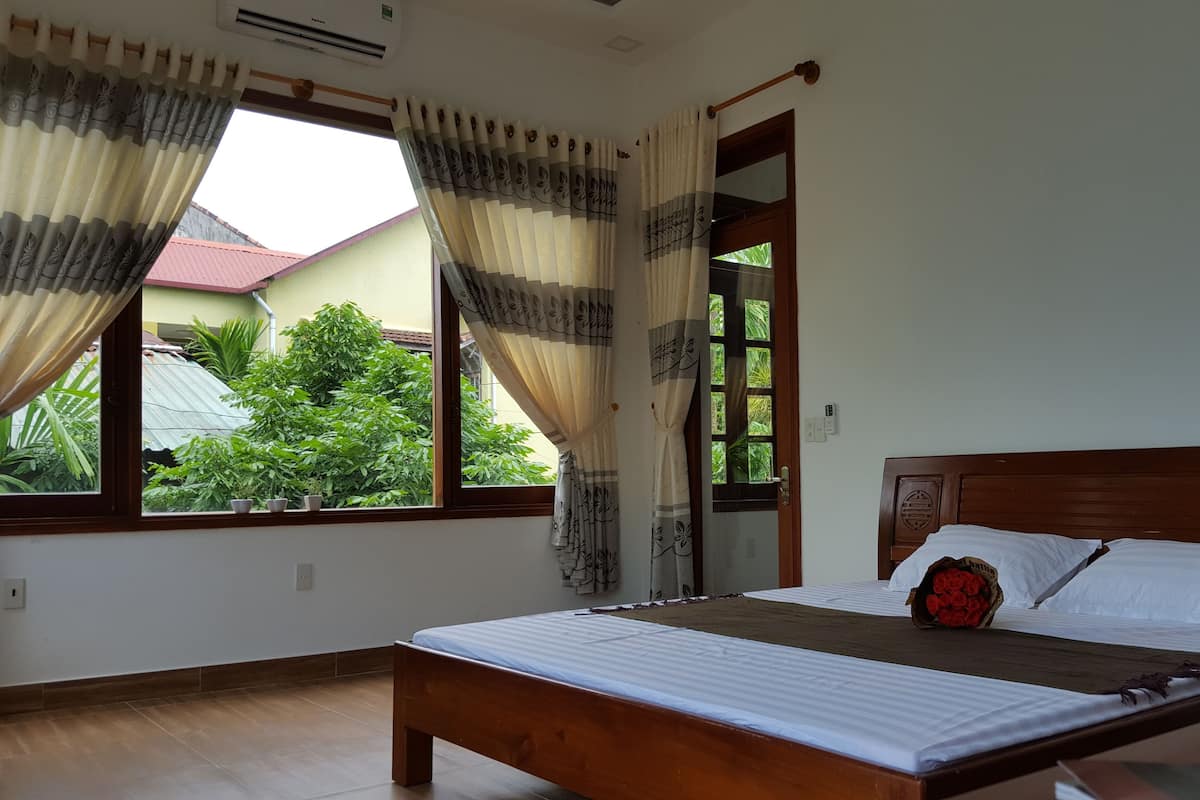 A bedroom with a wooden bed, a window with curtains, and a view of greenery outside.