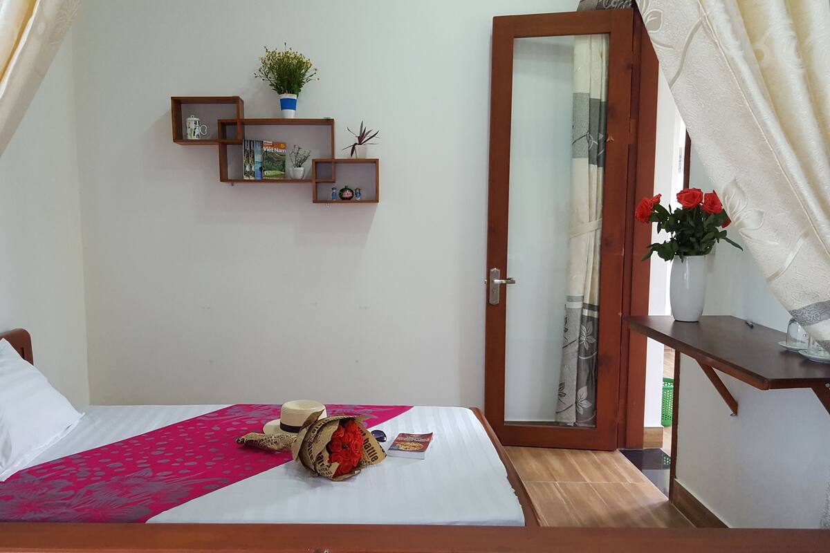 A neatly made bed with a pink bedspread, a wooden headboard, and a bedside table with a book and a hat. There is a small shelf with books and a potted plant on the wall.