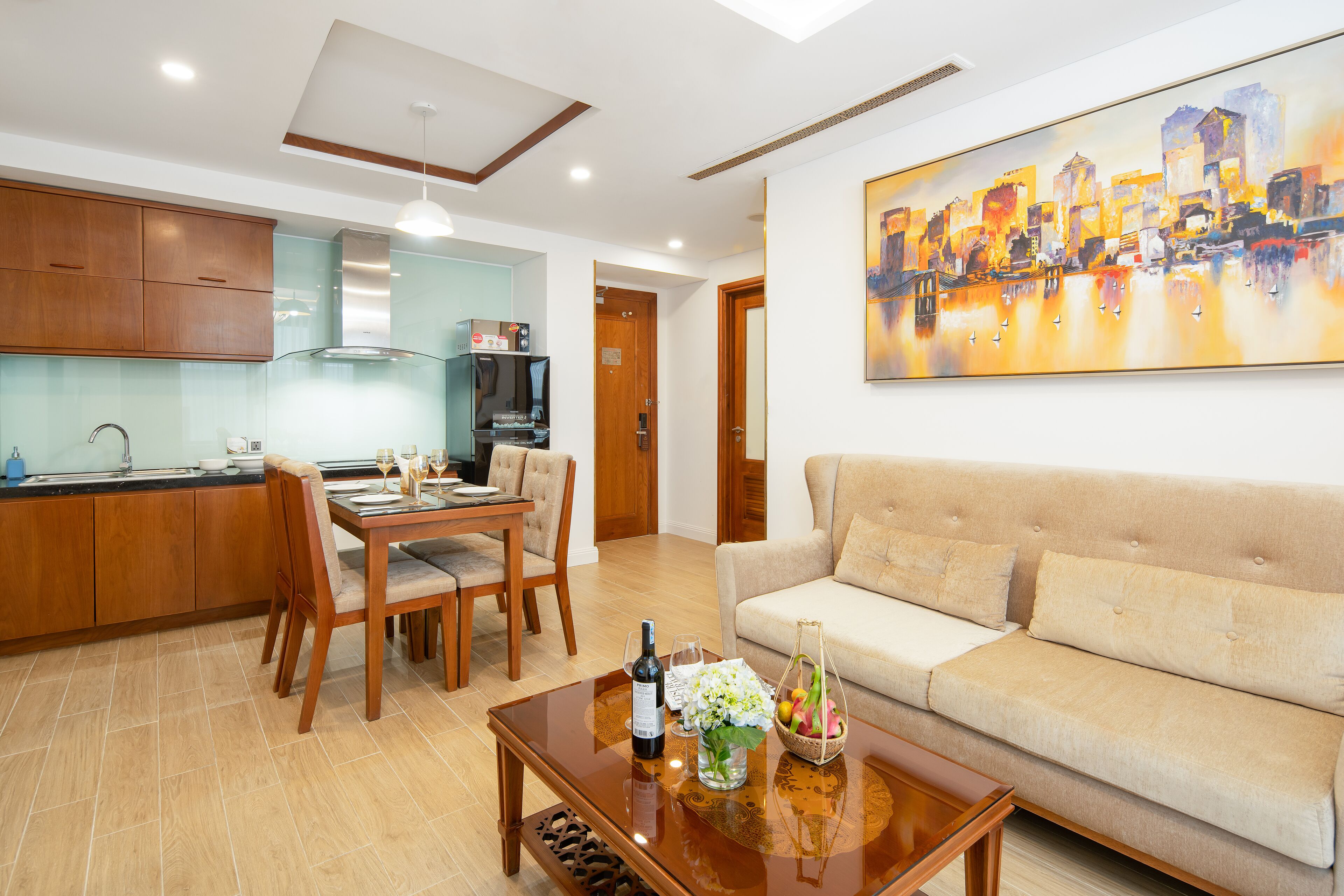 A modern living room with a sofa, a dining table with chairs, a coffee table with a bottle and flowers, and a kitchen in the background.