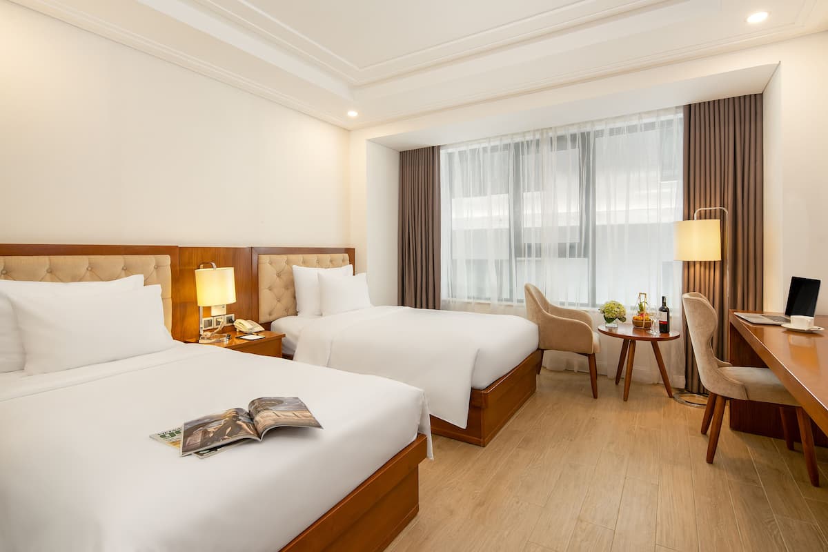 Une chambre d’hôtel avec deux lits, un bureau, une chaise, une lampe et une petite table sur laquelle se trouvent une bouteille et une plante.
