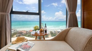 Een kamer met een groot raam dat uitkijkt op een strand, een bank, een klein tafeltje met een fles, glazen en een mand met fruit.