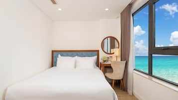 A hotel room with a bed, a desk with a chair, a round mirror, and a large window with a view of the ocean.