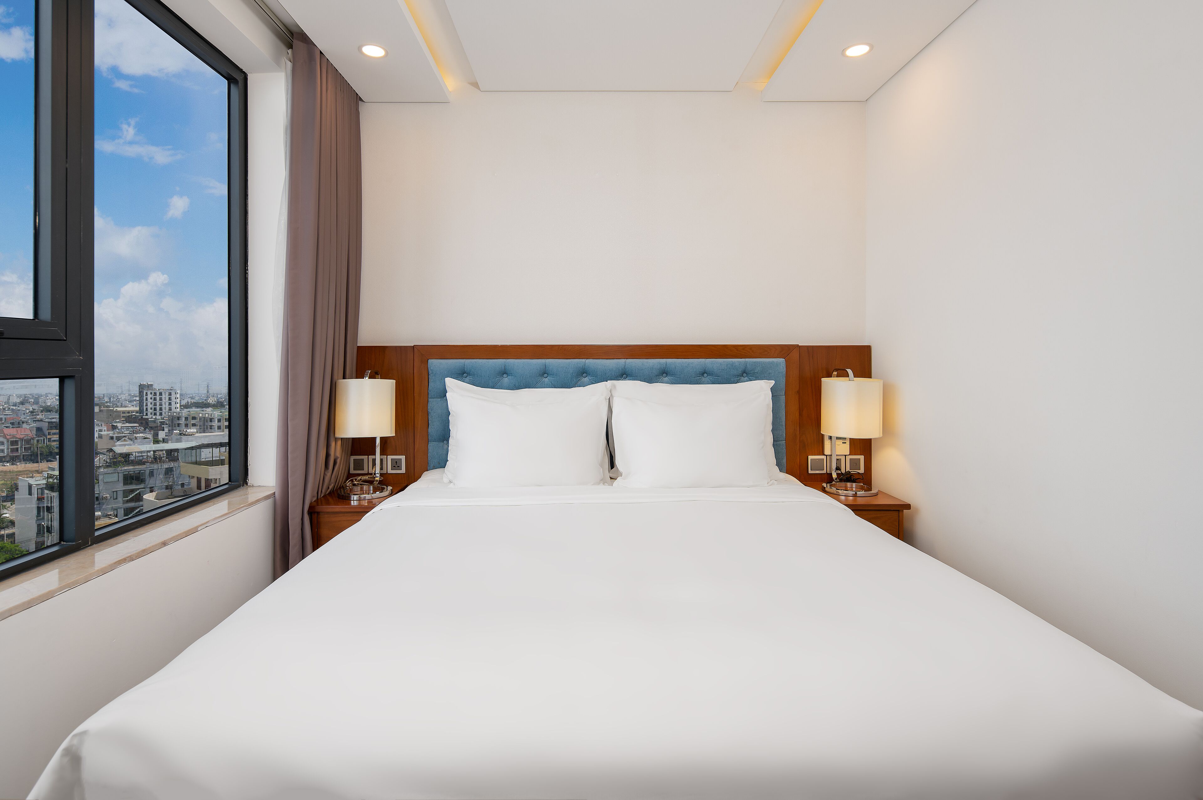 A hotel room with a large bed, two bedside lamps, a wooden headboard, and a view of the city through a large window.