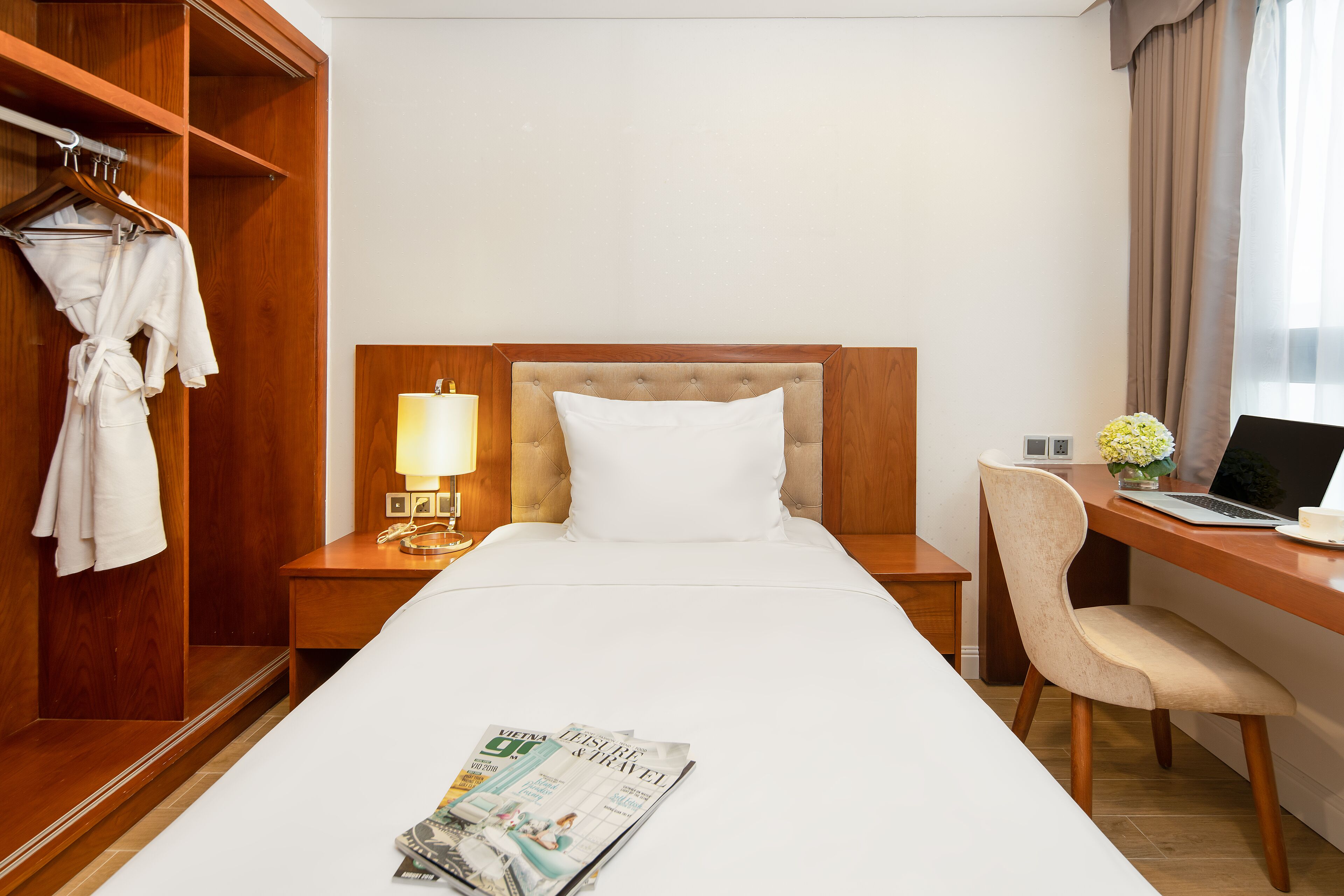 Una habitación de hotel con una cama, un escritorio con una computadora portátil, una silla, un armario y una revista sobre la cama.