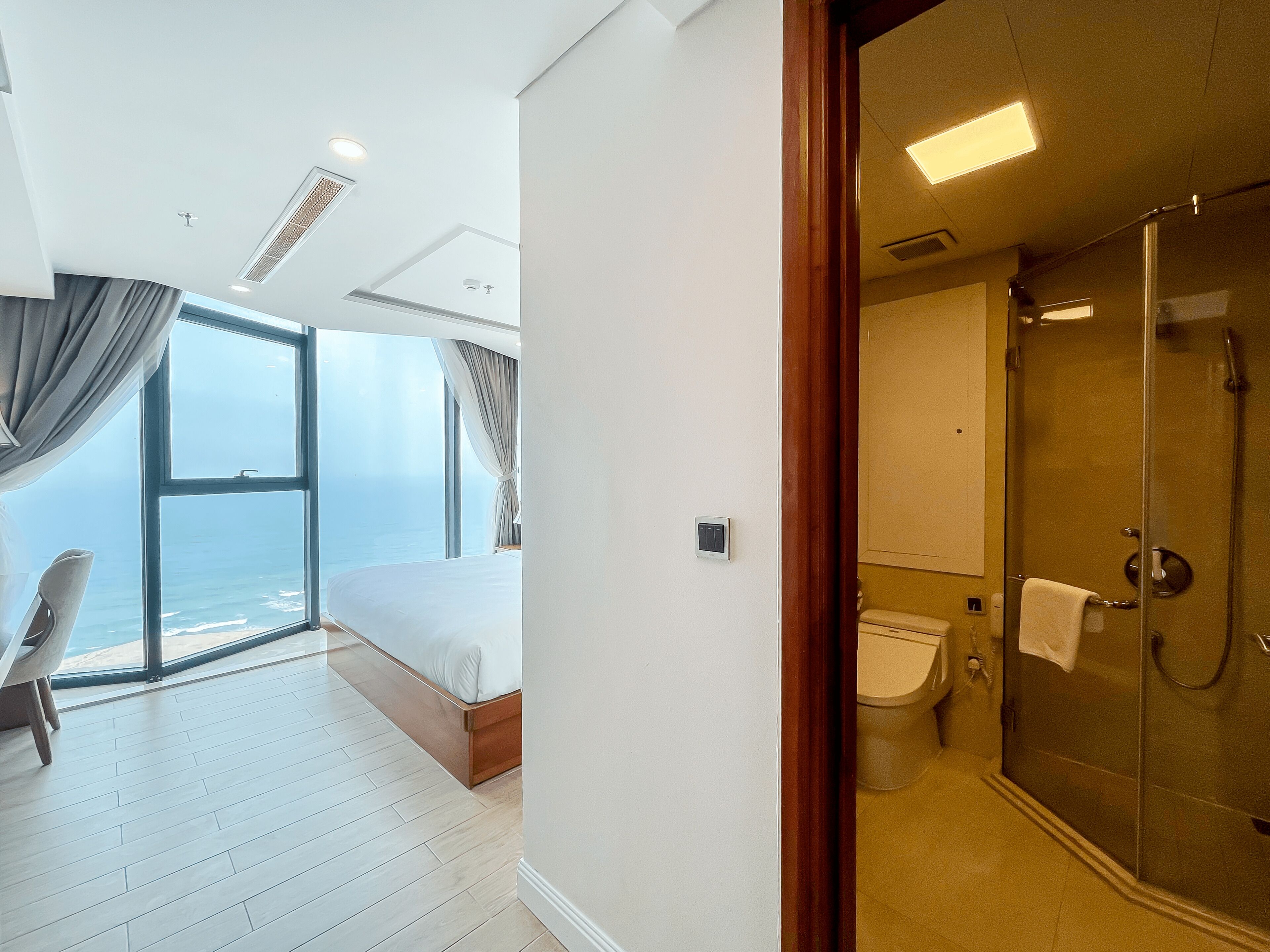 A modern hotel room with a large window offering a sea view, a separate bathroom with a glass shower, and a wooden floor.
