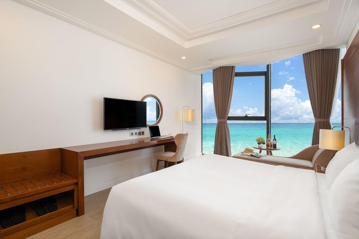 Une chambre d’hôtel avec un lit, un bureau avec un ordinateur portable, une télévision et une vue sur l’océan.