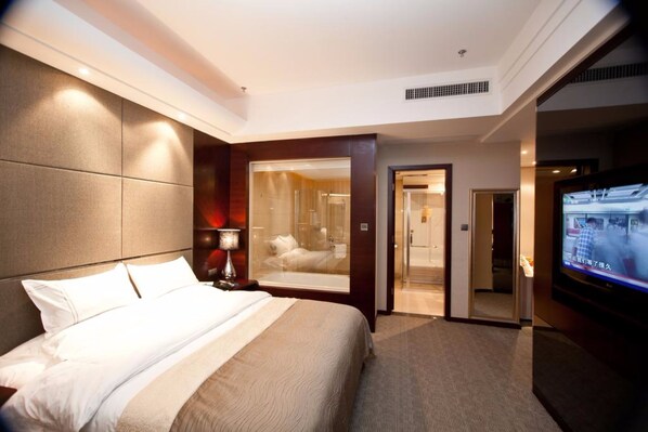 A hotel room with a large bed, a TV, a bathroom with a glass shower, and a door leading to another room.