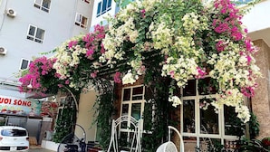 A building with a balcony covered in flowering plants, a white car parked in front, and a sign with Vietnamese text. - Hai Tien Plaza Hotel (Hoang Tien)