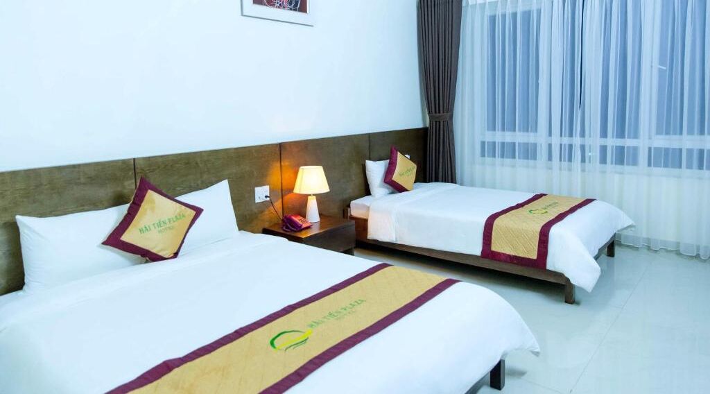 Une chambre d’hôtel avec deux lits, une table de chevet avec une lampe et une fenêtre avec des rideaux légers.