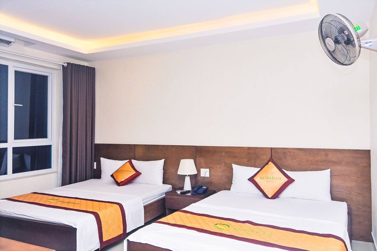 Ein Hotelzimmer mit zwei Betten, einem Nachttisch, einer Lampe, einem Ventilator und einem Fenster mit Vorhängen.