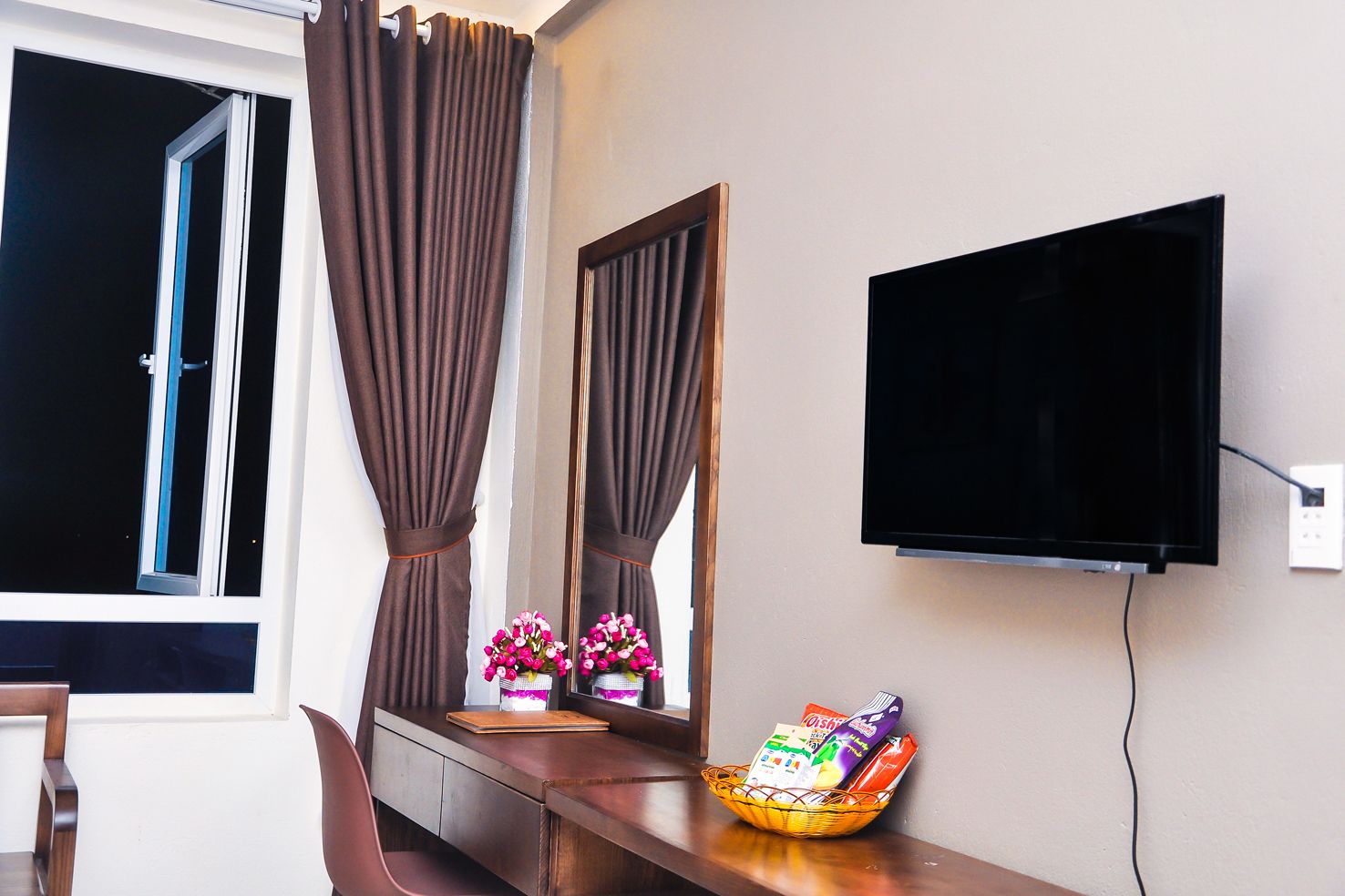 Een hotelkamer met een aan de muur bevestigde televisie, een bureau met een mand met snacks, een spiegel die de gordijnen weerkaatst en een raam met gordijnen.
