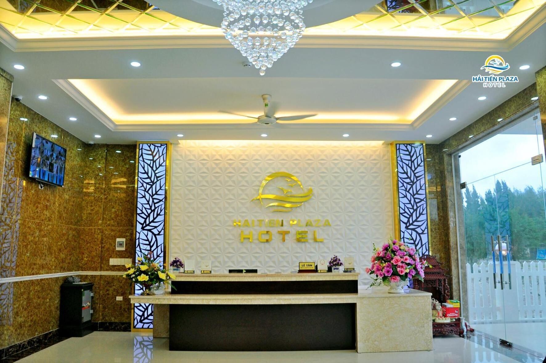 Un área de recepción de hotel con diseño moderno, que presenta un mostrador central con un logotipo, una araña y un amplio ventanal con vistas al exterior.