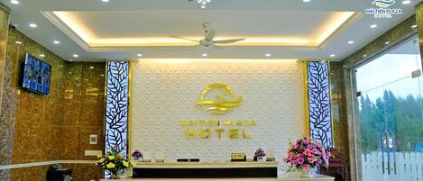 A hotel reception area with a modern design, featuring a central desk with a logo, a chandelier, and a large window offering a view of the outdoors.