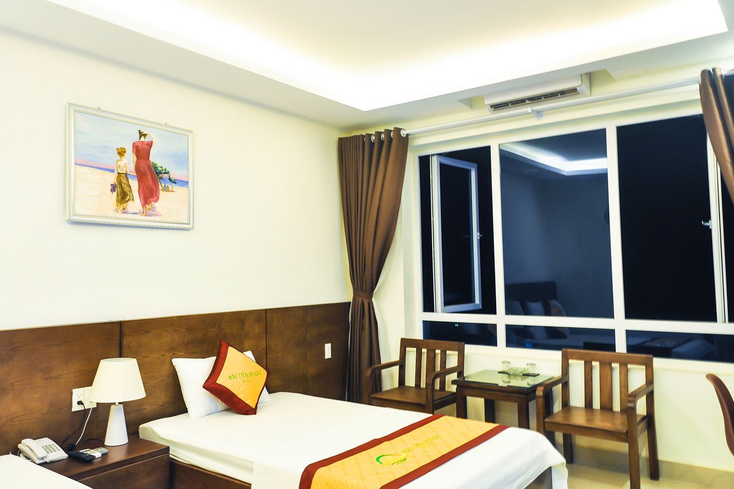 Habitación de hotel con cama, mesita de noche, lámpara y un cuadro en la pared. Se ve una ventana con cortinas y vistas al exterior.