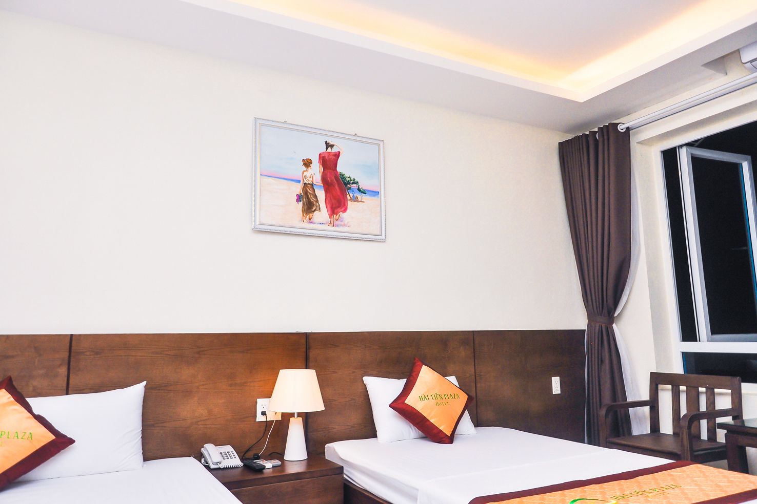 Een hotelkamer met twee bedden, een nachtkastje met een lamp, een schilderij aan de muur en een raam met gordijnen.