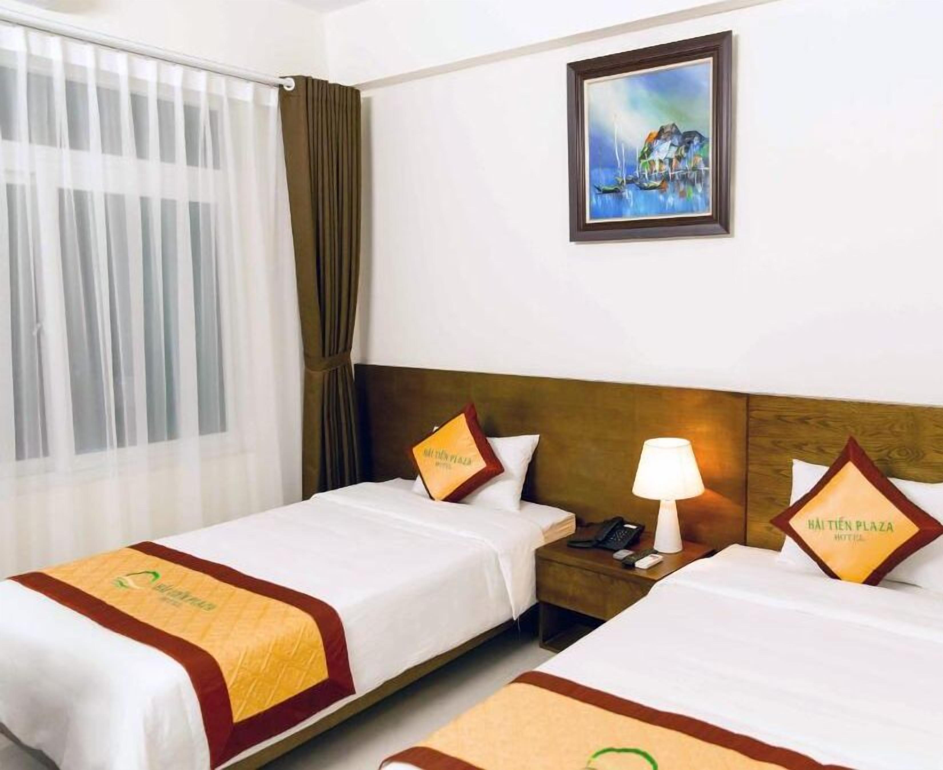 A hotel room with two beds, a painting on the wall, a window with curtains, and a nightstand with a lamp.