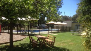 Un área exterior cercada con piscina, mesas de picnic y un pequeño edificio con sombrillas.