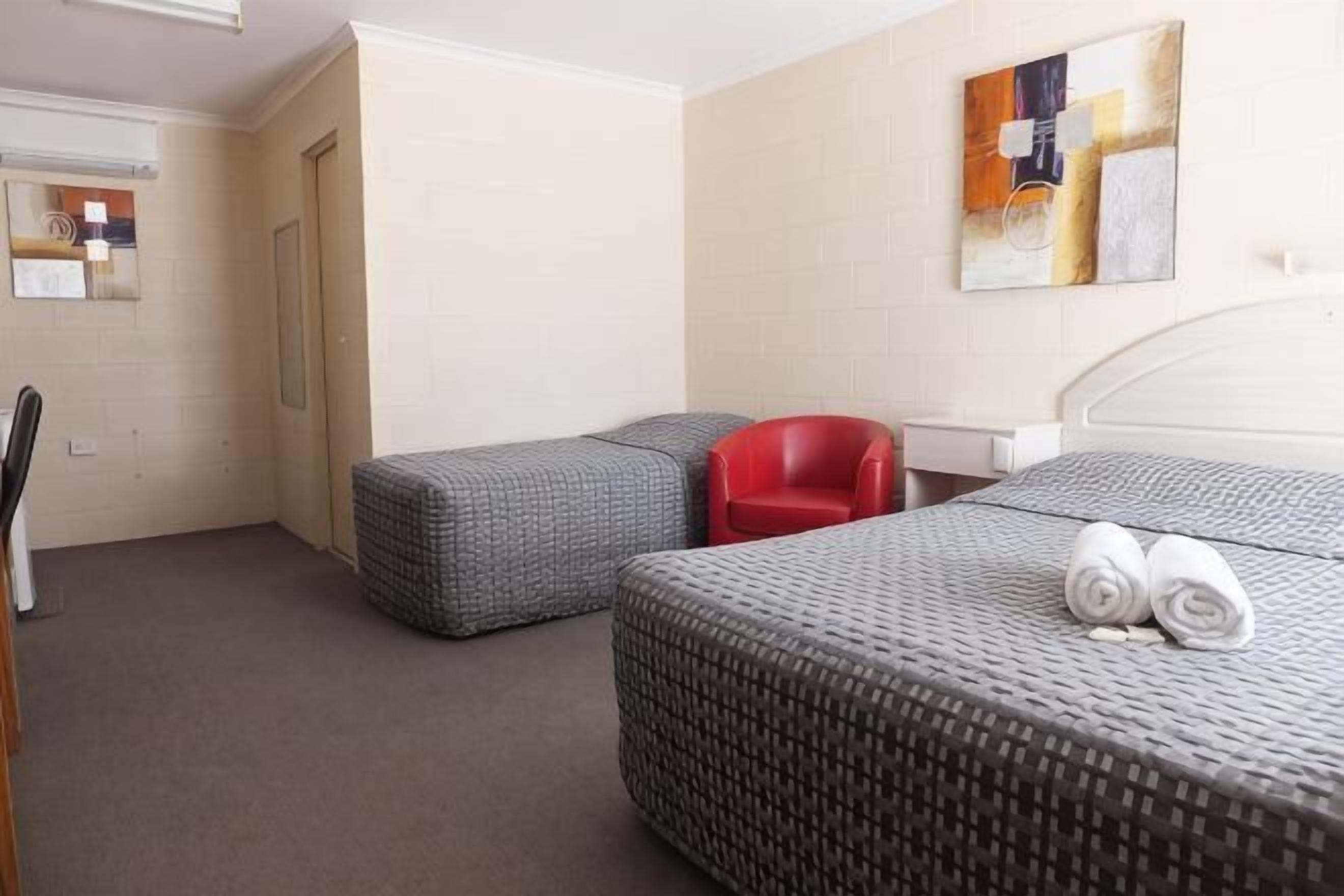 A hotel room with two beds, a red chair, a desk, and a painting on the wall.