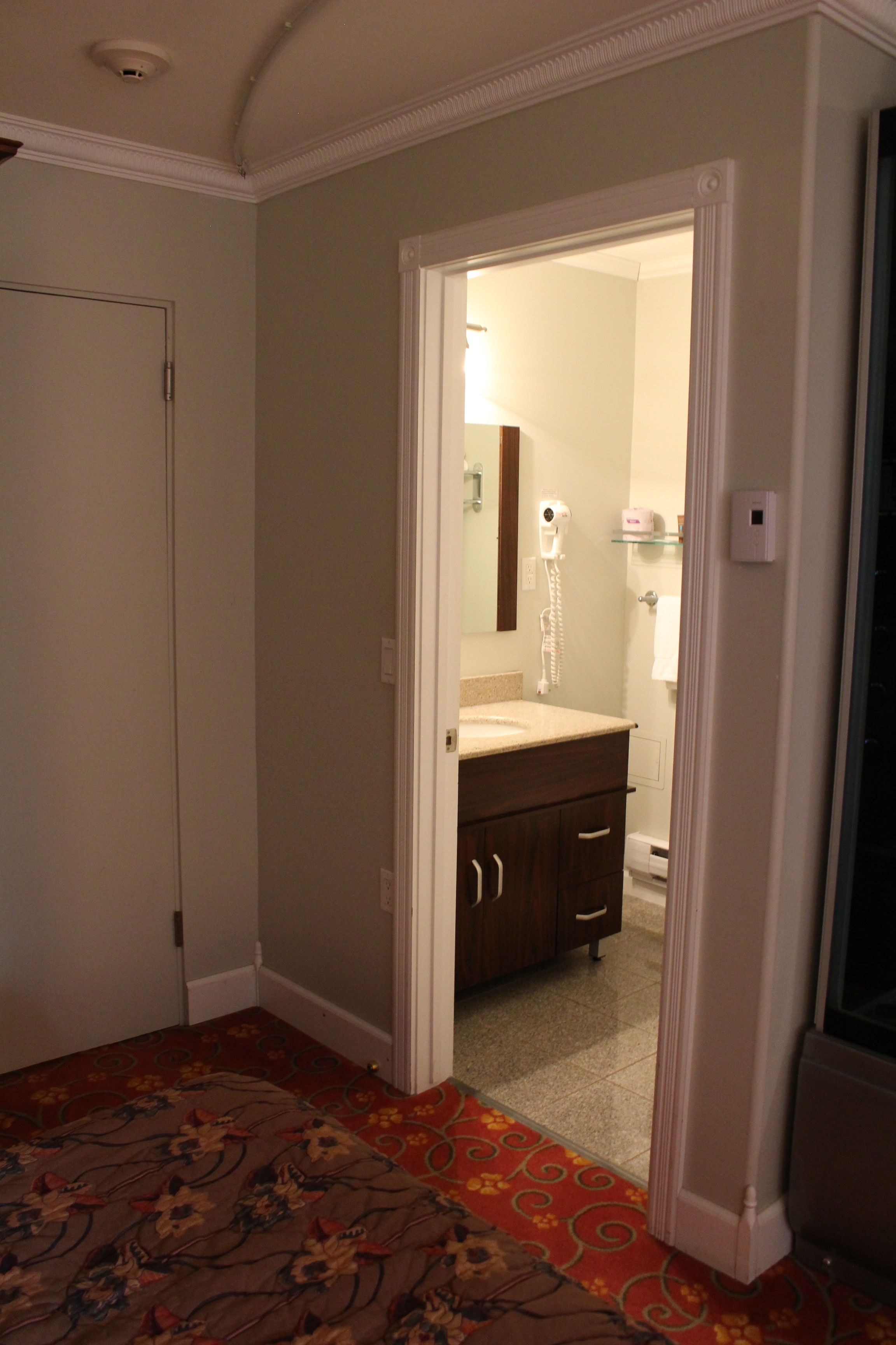 A hotel room with a carpeted floor, a bathroom with a sink and mirror, and a door leading to another room.