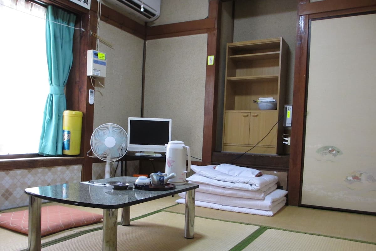 A hotel room with a tatami mat floor, a small table with a fan, a TV, a kettle, and neatly stacked towels.