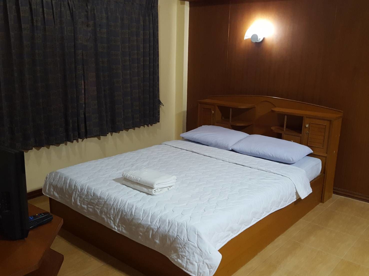 Una cama bien hecha con ropa blanca y almohadas, un cabecero de madera, una mesita de noche y una lámpara fijada a la pared.