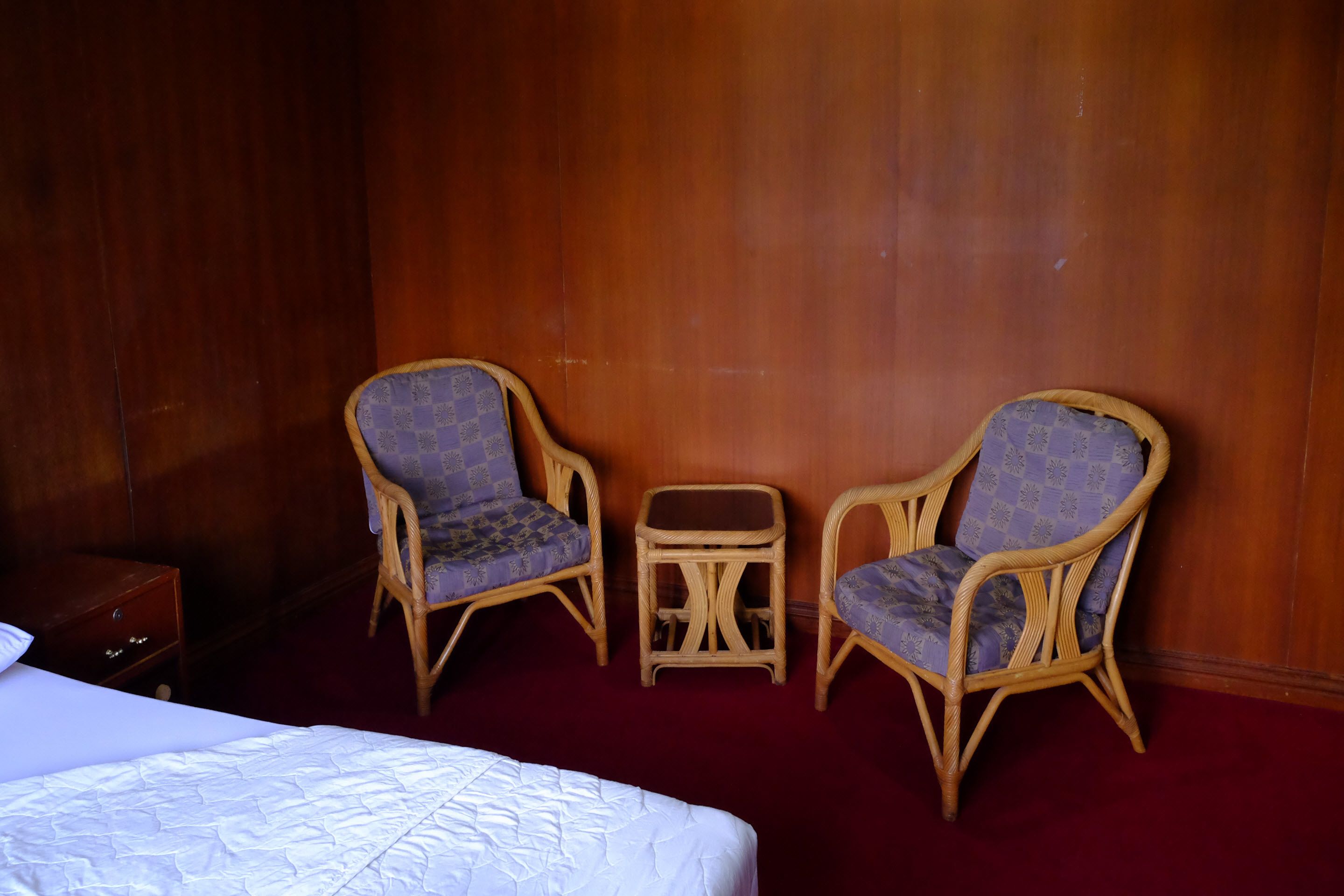 A hotel room with a bed, two wicker chairs, a small table, and a wooden wardrobe.