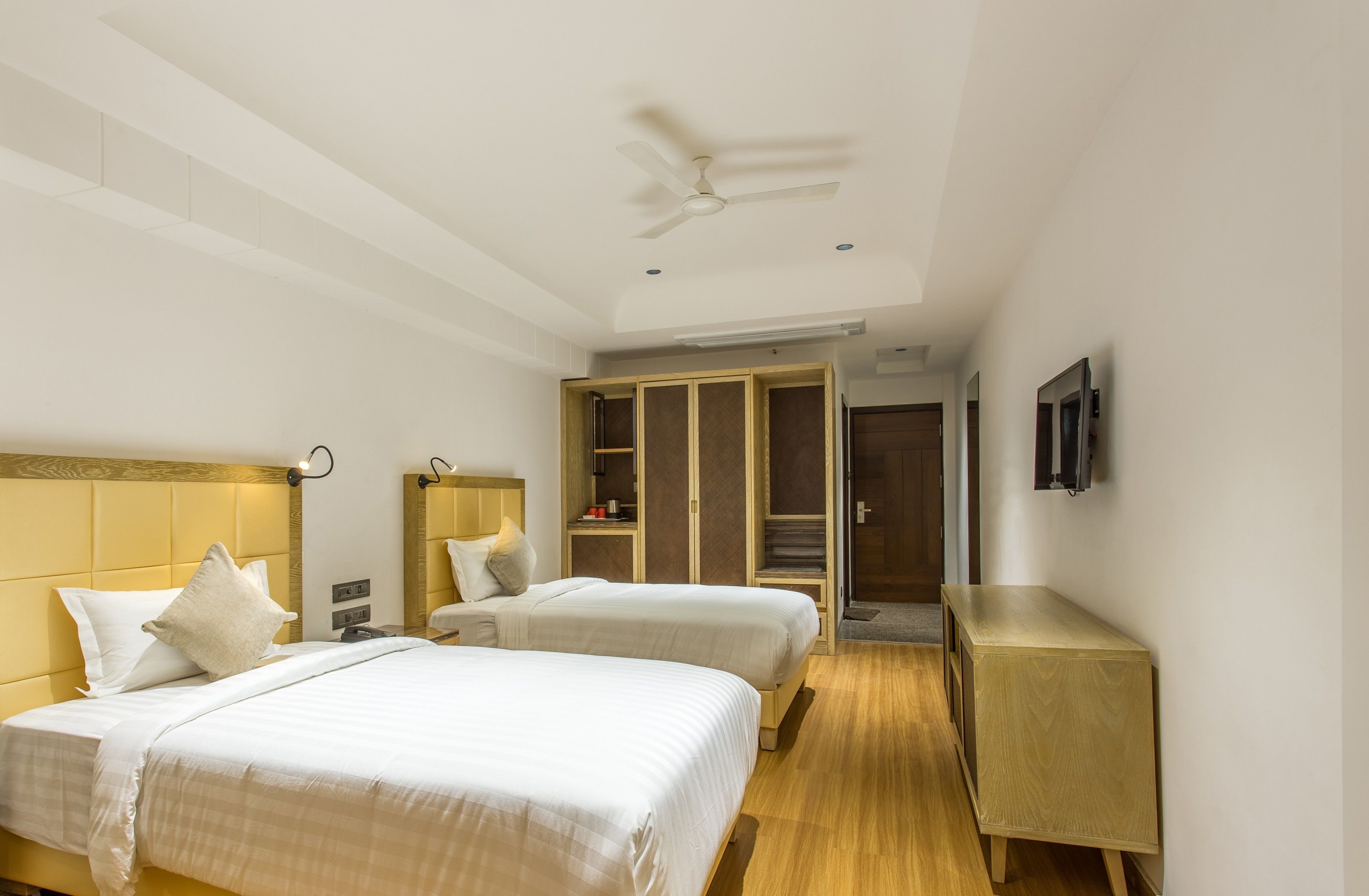 A hotel room with two beds, a ceiling fan, a wardrobe, a bedside table, and a TV mounted on the wall.