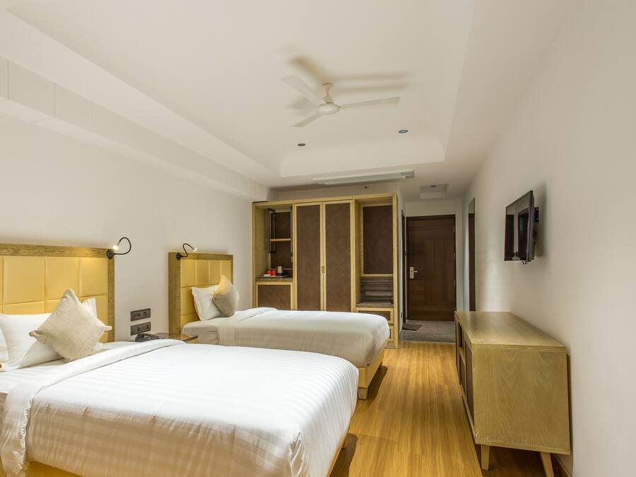 Quarto de hotel com duas camas, ventoinha no teto, guarda-roupa, mesa de cabeceira e televisão fixada na parede.
