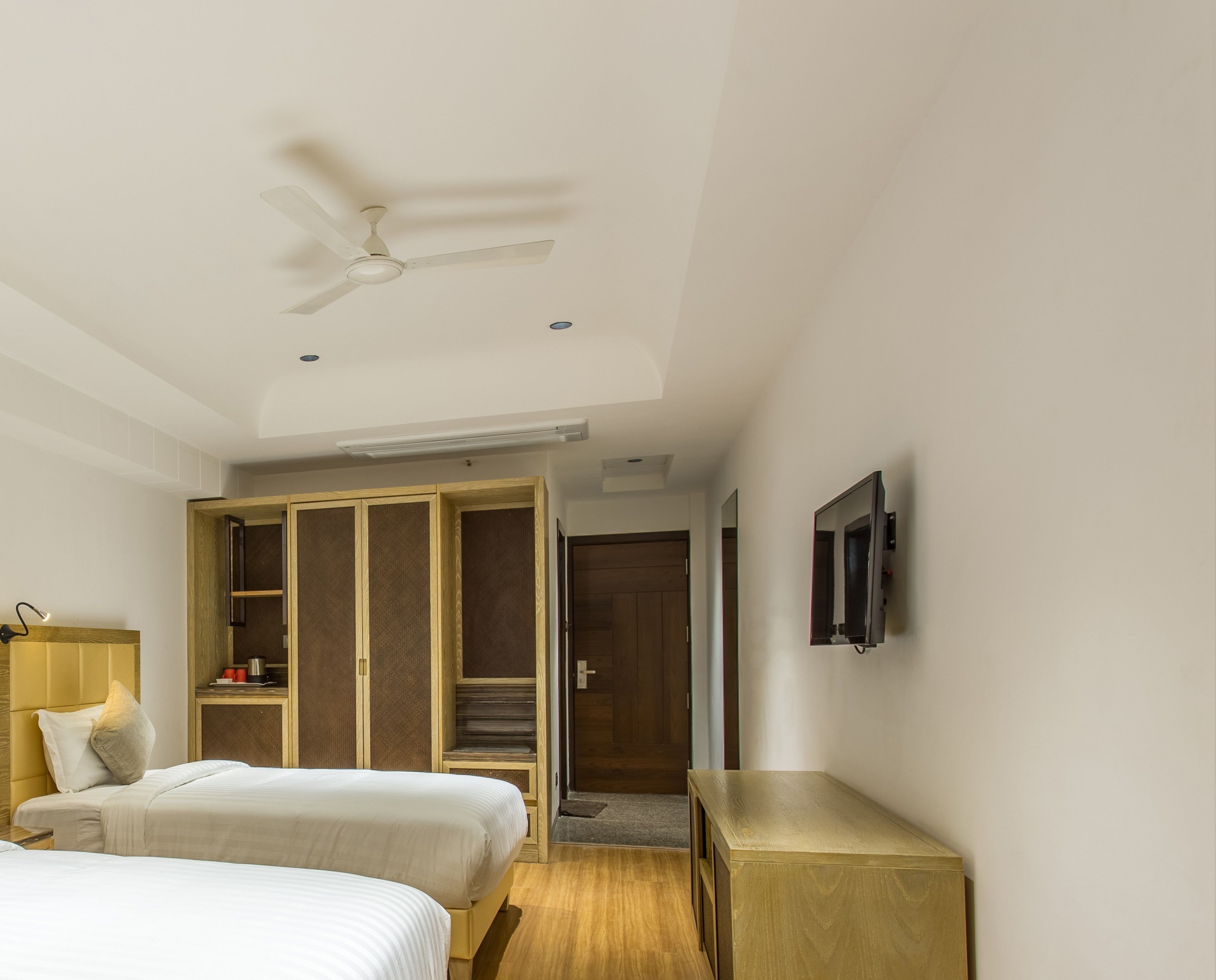 A hotel room with two beds, a ceiling fan, a TV mounted on the wall, and a wooden wardrobe.