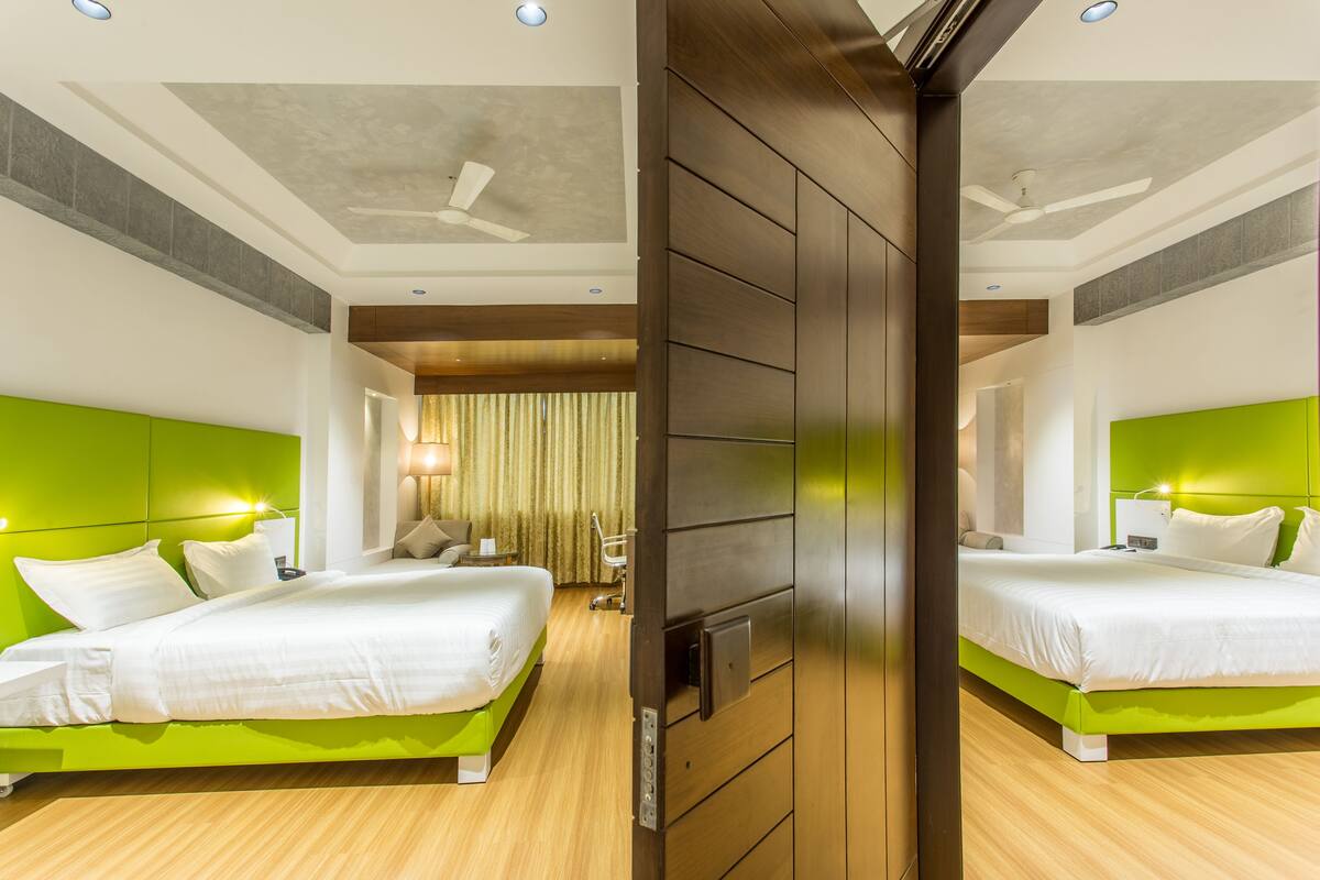 Habitación de hotel con dos camas, cabeceros verdes, suelo de madera, ventilador de techo y un baño visible al fondo.