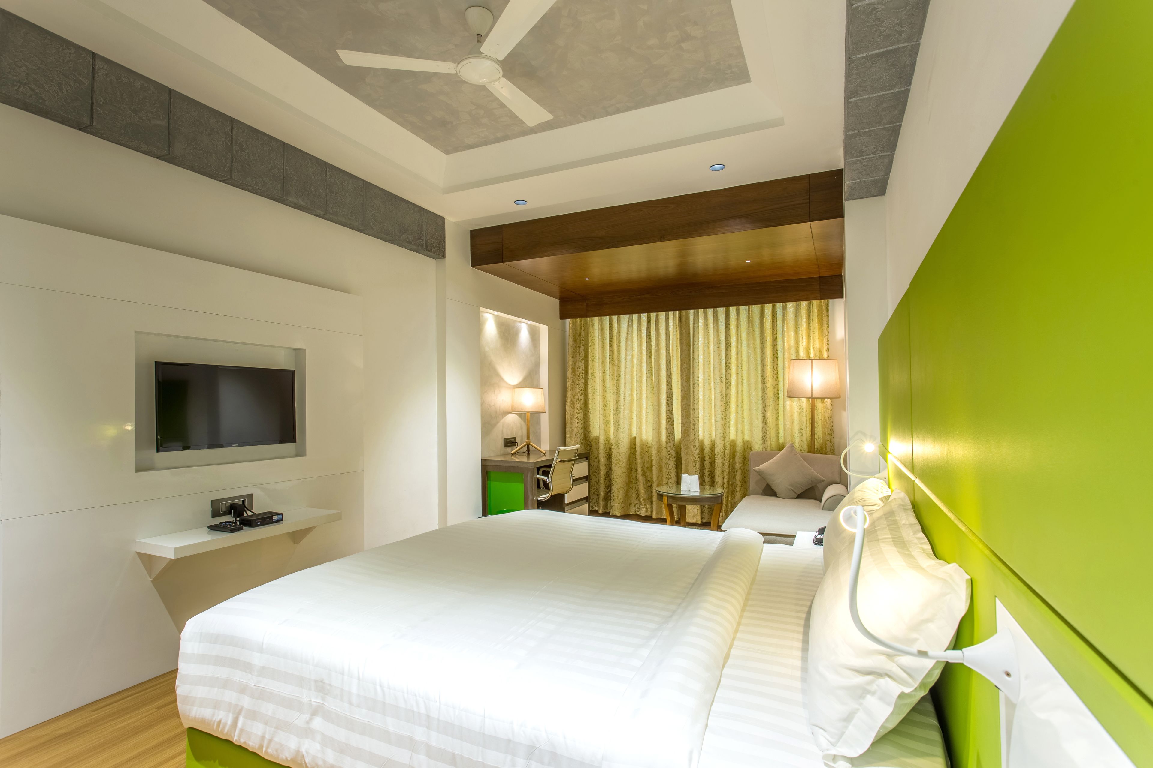 A modern hotel room with a large bed, a flat-screen TV, a desk with a lamp, and a green accent wall.
