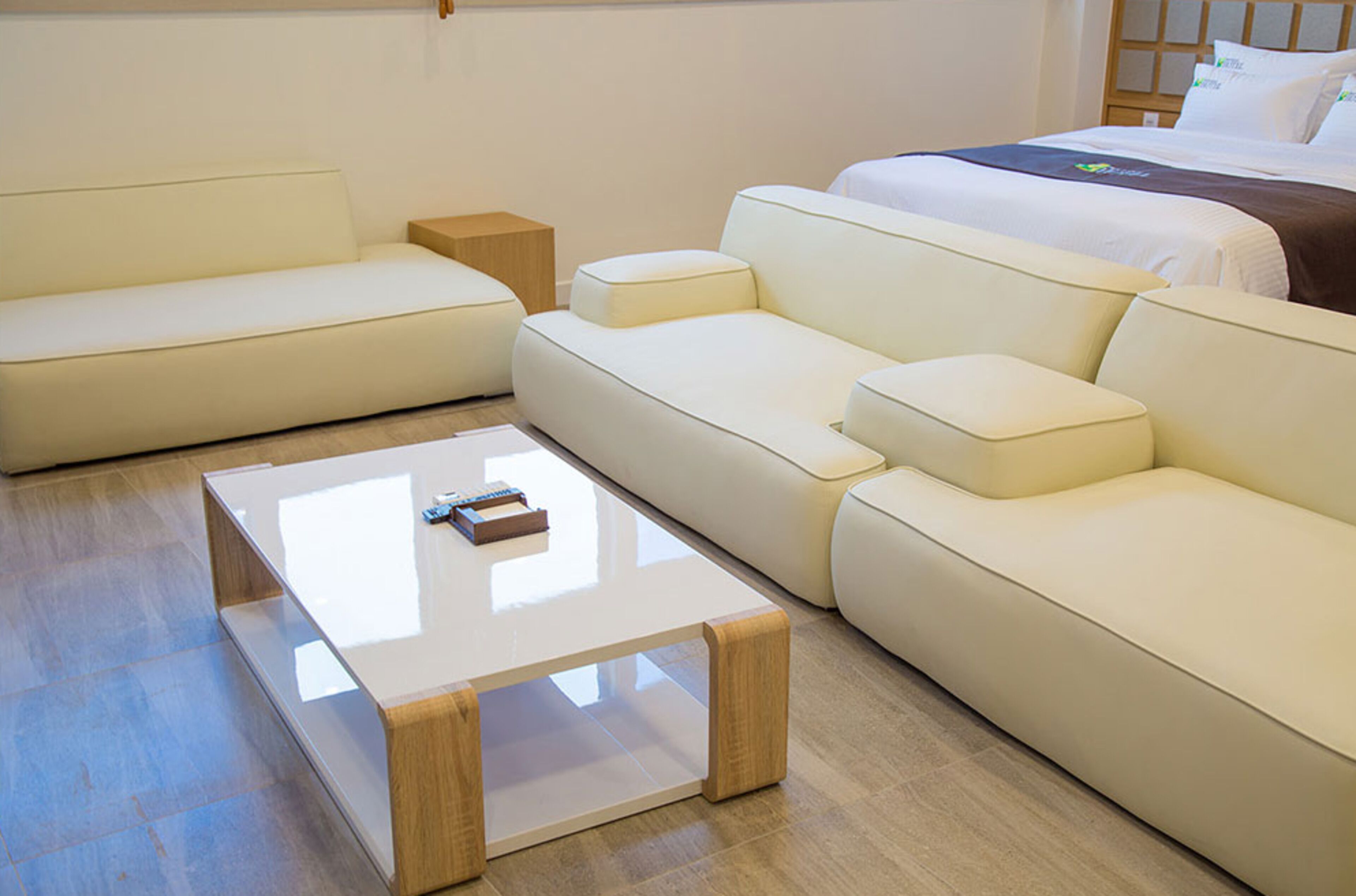 Una habitación de hotel moderna con un sofá color beige, una mesa de centro de madera y una cama con ropa blanca.